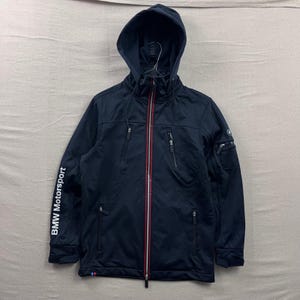May include: Navy blue hooded jacket with a full-length zipper and multiple pockets. The jacket features the text "BMW Motorsport" on the left sleeve and a red and white stripe detail on the zipper. A small logo is on the right sleeve.