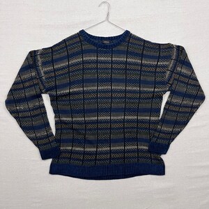 May include: A blue, tan, and white patterned sweater with a crew neck and long sleeves. The sweater has a geometric design with vertical and horizontal stripes. The sweater is hanging on a metal hanger against a white background.