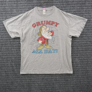 Könnte beinhalten: Ein hellgraues Kurzarm-T-Shirt mit einer Grafik der Figur Grumpy aus Schneewittchen. Der Text darüber lautet GRUMPY in Rot und der Text darunter lautet ALL DAY! in Blau. Im Hintergrund ist eine blasse Strichzeichnung der Figur zu sehen.