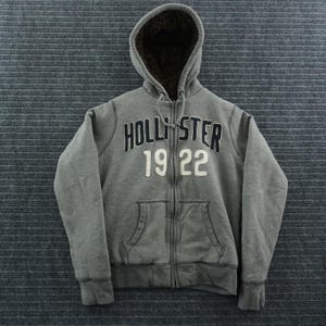 May include: A gray zip-up hoodie featuring a dark brown faux fur lining inside the hood. The front displays the text HOLLISTER in dark blue letters above the number 19 22 in white. It includes a full-length zipper, long sleeves, and two pockets.