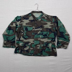 Puede incluir: Una chaqueta con estampado de camuflaje con múltiples bolsillos delanteros y puños abotonados. La chaqueta presenta una combinación de colores verde, marrón y negro, y está colgada de una percha de metal. La chaqueta tiene cuello.