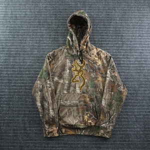 May include: A long-sleeve hooded sweatshirt featuring a realistic forest camouflage pattern in shades of brown, green, and tan. The center of the chest displays a large, embroidered gold-colored Browning buckmark logo above a front kangaroo pocket.
