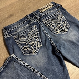 以下が含まれることがあります： 装飾的なステッチと装飾されたバックポケットが付いた青いデニムジーンズ。ジーンズは、バックウエストバンドにRock Revivalのラベルが付いています。ポケットには、詳細な刺繍デザインがあります。