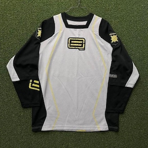 Puede incluir: Una camiseta de manga larga con cuerpo blanco y mangas negras. La camiseta presenta un cuello cuadrado y detalles amarillos. Un logotipo amarillo está en el pecho y un emblema en forma de estrella en cada hombro.