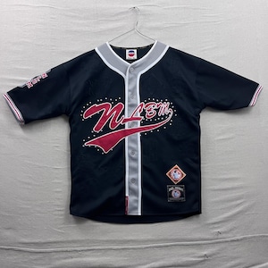 Peut inclure: Maillot de baseball noir avec le texte "NMBM" en script rouge et blanc sur le devant. Le maillot a un col gris et un devant boutonné, avec une bordure rouge et blanche sur les manches. Un petit écusson est visible en bas à droite.