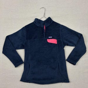 May include: Navy blue fleece pullover with a high collar and a pink chest pocket. The pullover has a button closure at the neck and a small brand patch on the chest pocket. The long-sleeved top is hanging on a metal hanger.