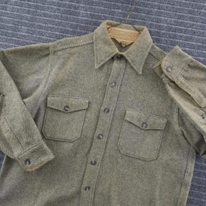 May include: A long-sleeve button-down shirt in a textured olive green fabric. The shirt features a pointed collar, two chest pockets with buttoned flaps, and matching buttons down the front placket and on the cuffs. The interior collar lining is tan.