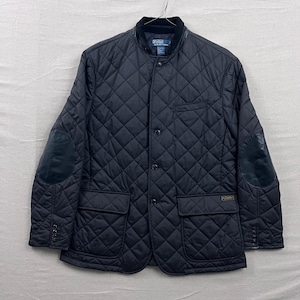 May include: Black quilted jacket with a corduroy collar, leather elbow patches, and two front pockets. The jacket has a button-up front and a small logo patch on the pocket. The jacket is hanging on a metal hanger.