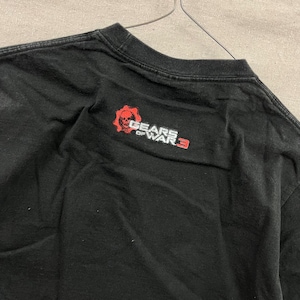 Puede incluir: Camiseta negra con el logo de Gears of War 3. El logo incluye una calavera roja y las palabras "GEARS OF WAR 3" en blanco. La camiseta está hecha de un material suave.