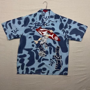 May include: Light blue button-up shirt with a dark blue abstract pattern. The shirt features a cartoon surfer graphic holding a red and white surfboard. The words "Wave Crasher" are printed on the lower right side of the shirt.
