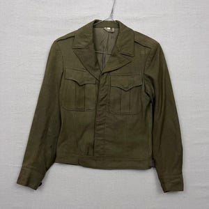 Puede incluir: Chaqueta verde oliva con dos bolsillos en el pecho y dos bolsillos inferiores. La chaqueta tiene cuello con solapas y puños abotonados. Cuelga de una percha metálica. La chaqueta parece estar hecha de una tela duradera.