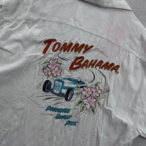 Puede incluir: Camisa abotonada de color blanquecino con un diseño bordado colorido en la espalda. El diseño presenta las palabras "Tommy Bahama" en rojo y un coche de carreras azul claro con flores. También se incluye el texto "Paradise Grand Prix".