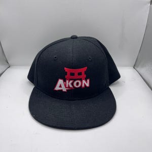 Puede incluir: Gorra de béisbol negra con visera plana. La gorra presenta la palabra "AKON" en letras blancas con un gráfico rojo encima. La gorra está hecha de un material resistente y tiene un ajuste estructurado.