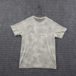 May include: A short-sleeve crew neck t-shirt in a light beige color featuring a subtle, tonal camouflage pattern created by horizontal lines. The garment has a seamless construction with raglan sleeves and a ribbed neckline, presented on a hanger.
