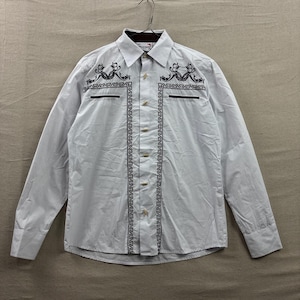 Peut inclure: Une chemise blanche à manches longues boutonnée avec des motifs de volutes noirs brodés sur la poitrine et des bordures géométriques verticales entourant la patte de boutonnage centrale. Elle présente deux poches poitrine noires.
