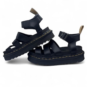 May include: A pair of black leather sandals with multiple straps and a thick, ridged sole. The sandals have adjustable ankle straps with buckles and are designed for comfort and style. The stitching is a contrasting yellow color.