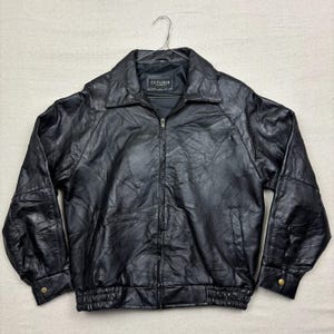 Könnte beinhalten: Schwarze Lederjacke mit Patchwork-Design. Die Jacke hat einen Reißverschluss, einen Kragen und zwei Vordertaschen. Die Ärmel haben geknöpfte Manschetten und die Taille hat ein elastisches Band. Die Jacke hängt an einem Metallbügel.