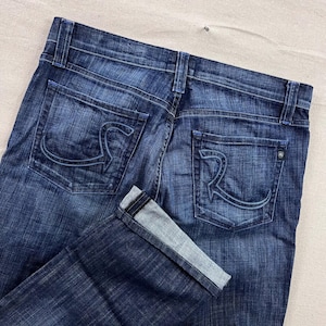 May include: Dark wash denim jeans with a classic five-pocket design. The back pockets feature a stylized "R" design. The jeans have a folded cuff, and the stitching is a contrasting light blue color.