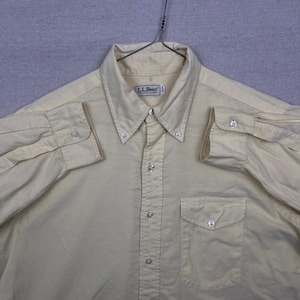 May include: A light yellow button-down shirt with long sleeves, a button-down collar, and a chest pocket. The shirt has the brand label "L.L.Bean" on the collar. The shirt is hanging on a metal hanger.
