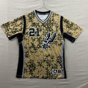 Peut inclure: Un maillot de basket-ball à manches courtes avec un motif de camouflage numérique dans les tons de beige, de vert et de noir. Le maillot porte le numéro "21" en noir, le logo des Spurs et le logo Adidas. Le maillot a un col en V.