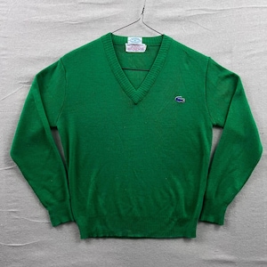 Op de afbeelding: Een groene V-hals trui met lange mouwen. De trui heeft een klein blauw embleem op de borst en een label bij de halslijn. De trui hangt aan een draadhanger.