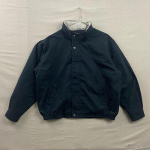 May include: A dark navy blue jacket featuring a high collar with a light beige interior lining. The jacket includes a front zipper closure concealed by a snap-button placket, long sleeves with cuffs, and an elasticized hemline at the waist.