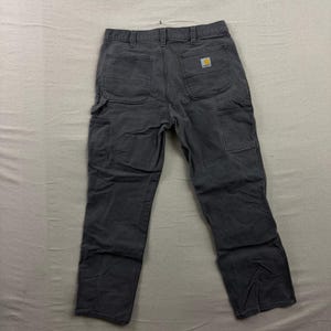 May include: A pair of charcoal gray work pants featuring a multi-pocket design, including two large back pockets and additional utility pockets on the legs. A small rectangular Carhartt brand patch is sewn onto the right back pocket.