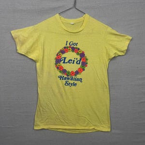May include: Yellow t-shirt with the text "I Got Lei'd Hawaiian Style" in blue and a floral wreath design in orange, purple, and green. The shirt is hanging on a metal hanger.