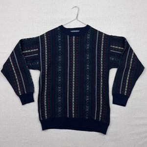 May include: A navy blue sweater with a striped pattern in white, burgundy, and green. The sweater has a crew neck and long sleeves with navy blue cuffs. The garment is hanging on a silver hanger against a white background.