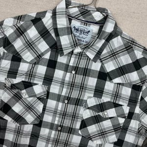 Puede incluir: Camisa a cuadros negros, blancos y grises con dos bolsillos en el pecho. La camisa tiene cuello y una etiqueta con un logotipo de caballo. La camisa está hecha de una tela ligera.