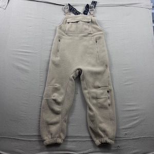 May include: Beige fleece overalls with black and white straps. The overalls feature a chest pocket, two zipped pockets, and reinforced knees. The straps have the text "EB W" printed on them. The overall's legs have a pocket on each leg.