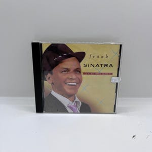 Puede incluir: Un CD de Frank Sinatra de la serie Collector's. La portada muestra un retrato sonriente de Sinatra con sombrero y traje. El fondo es de color beige claro con destellos dorados. La caja del CD es negra.