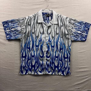 May include: White button-down shirt with a blue and gray flame design. The short-sleeved shirt has a collared neckline and is hanging on a metal hanger. The shirt's design features blue flames with gray outlines.