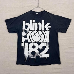 May include: Black t-shirt with the band name "blink 182" in large white letters. The design includes a smiley face with crossed-out eyes and a distressed, grunge-style aesthetic. The shirt is hanging on a metal hanger.