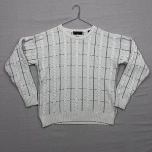 Puede incluir: Suéter de punto trenzado blanco con un patrón de cuadrícula gris. El suéter tiene cuello redondo y mangas largas. Se muestra colgado en una percha de metal.
