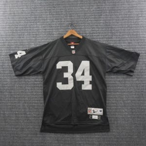 May include: A black short-sleeved football jersey featuring the number 34 in white on the front and sleeves. The jersey includes an NFL logo at the neckline, a size M tag at the bottom, and a small logo patch on the lower front. It is displayed on a hanger.