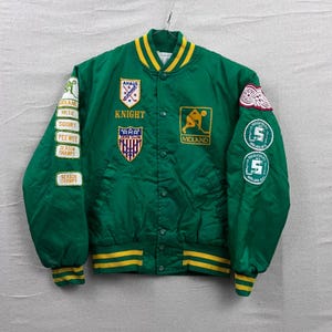 Op de afbeelding: Groene satijnen varsity jack met gele en witte accenten. Voorzien van patches met tekst als "Knight", "Midland" en "Season Champs". De jas heeft een knoopsluiting en geribde manchetten.