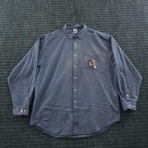 Könnte beinhalten: Ein langärmeliges Button-Down-Hemd aus blauem Stoff. Das Hemd verfügt über einen spitzen Kragen, eine einzelne Brusttasche mit einer Stickerei von Bugs Bunny und dem Tasmanischen Teufel sowie ein kleines rechteckiges Etikett am unteren Saum.