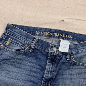 May include: Close-up of blue denim jeans with the text "NAUTICA JEANS CO." across the top. The jeans have a classic five-pocket design, a button and zipper closure, and a yellow logo on the front pocket.