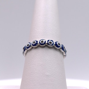 May include: A silver ring with a repeating pattern of blue and white evil eye designs. Each eye has a blue background, a white iris, and a black pupil. The ring is shown on a white surface.