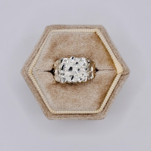 May include: A silver-colored nugget ring with a textured surface, set in a beige hexagonal ring box. The ring has a chunky, irregular design, with a mix of shiny and matte areas. The ring box has a soft, velvet-like interior.