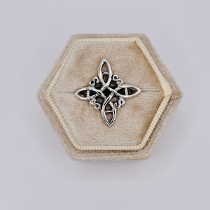 May include: A silver-colored Celtic knot pendant displayed in a beige velvet jewelry box. The pendant features an intricate, interwoven design. The box is hexagonal with a cream-colored trim.