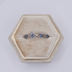 May include: A silver ring with a repeating evil eye design. Each eye features a blue iris and a white outline. The ring is displayed in a beige hexagonal jewelry box.