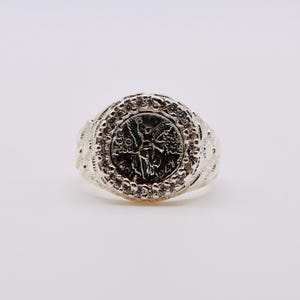 May include: A silver-toned ring featuring a circular centerpiece that resembles a coin with an embossed winged figure and the year 1947. The coin is surrounded by a border of small, clear, sparkling stones, and the band has a textured, patterned finish.