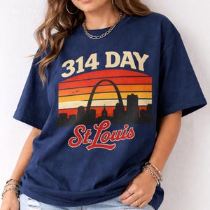 May include: Navy blue t-shirt featuring a retro sunset design with the text "314 DAY" and "St. Louis". The design includes the Gateway Arch and a city skyline in silhouette.