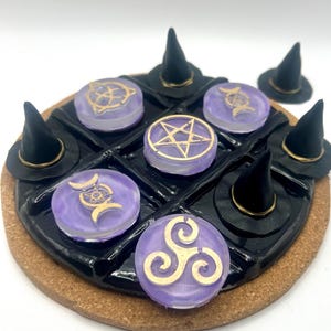 May include: A decorative coaster featuring a black grid design with purple and gold accents. The coaster has small black witch hats and circular purple pieces with gold symbols. The base is a brown cork material.