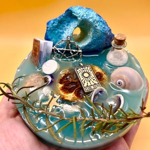 May include: A handcrafted resin art piece featuring a turquoise base with embedded seashells, a dried orange slice, and a pendant with "THE SUN" text. A small book, a pentagram charm, and a blue rock formation are also included.