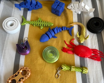 Könnte beinhalten: Eine Sammlung farbenfroher 3D-gedruckter Objekte, darunter blaue und weiße Haie, ein blauer Würfel, ein grünes Fischskelett, eine rote Krabbe und ein goldfarbenes Kettenglied-Design. Weitere Artikel sind eine grüne Spirale, ein lila Schlüsselanhänger und eine schwarze Scheibe.