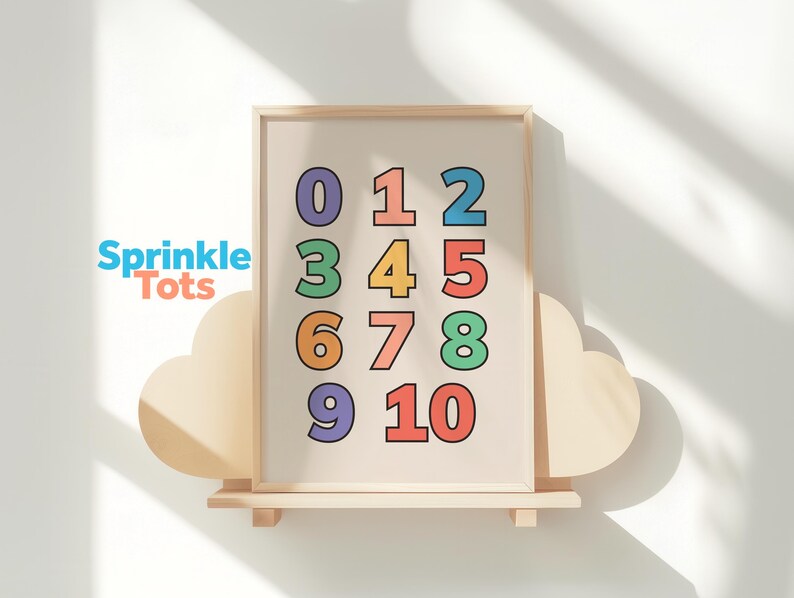 May include: A framed print featuring the numbers 0 through 10 in various colors. The numbers are displayed on a cream-colored background. The print is displayed on a wooden shelf with cloud-shaped accents. The brand name "Sprinkle Tots" is visible.