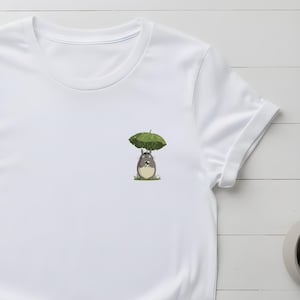 May include: White t-shirt featuring a small graphic of a cartoon character under a green umbrella. The character is gray and white, sitting on green grass. The shirt has short sleeves and a crew neck.
